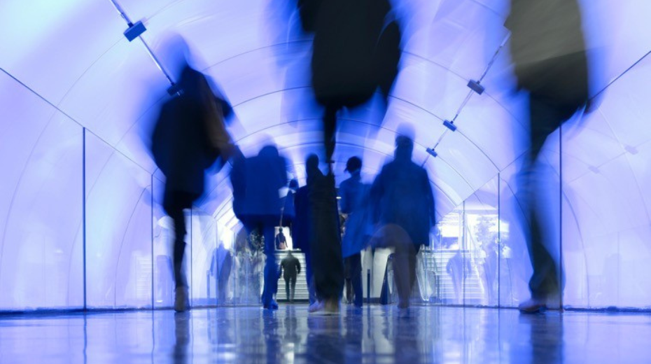Blurred silhouettes of people walking through a futuristic blue-lit tunnel.
